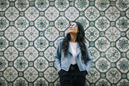 Portrait of young woman wearing light blue leather jacket and sunglasses in front of Azulejo wall, Lisbon, Portugal - DCRF00029