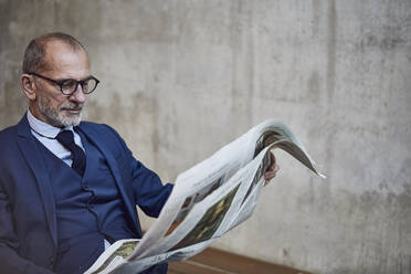 Mature businessman reading newspaper in front of concrete wall - MCF00595