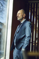 Senior businessman standing in his sustainable office, looking out of window - MCF00624