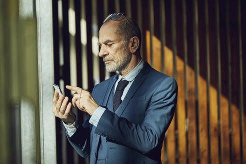 Senior businessman standing in his sustainable office, using smartphone - MCF00628