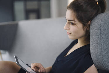 Junge Geschäftsfrau sitzt mit digitalem Tablet auf der Couch und schaut in die Ferne - KNSF07627
