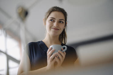Porträt einer lächelnden jungen Frau mit einer Tasse Kaffee im Büro - KNSF07632
