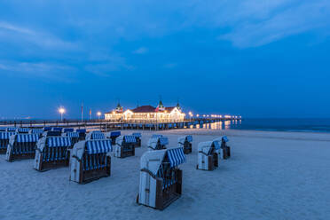 Germany, Mecklenburg-Western Pomerania, Heringsdorf, Hooded beach chairs on sandy coastal beach at dusk with illuminated pier in background - WDF05836