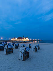 Germany, Mecklenburg-Western Pomerania, Heringsdorf, Hooded beach chairs on sandy coastal beach at dusk with illuminated pier in background - WDF05837