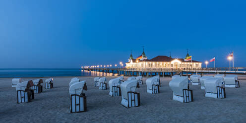 Germany, Mecklenburg-Western Pomerania, Heringsdorf, Hooded beach chairs on sandy coastal beach at dusk with illuminated pier in background - WDF05846