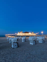 Germany, Mecklenburg-Western Pomerania, Heringsdorf, Hooded beach chairs on sandy coastal beach at dusk with illuminated pier in background - WDF05847