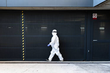Female scientist wearing protective suit and mask and walking in front of a wall - ERRF02744