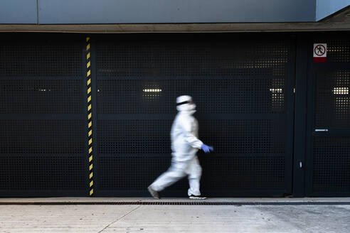 Female scientist wearing protective suit and mask and walking in front of a wall - ERRF02745