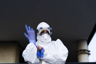 Wissenschaftlerin trägt Schutzanzug und -maske und zieht Schutzhandschuhe an - ERRF02747