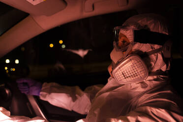 Senior woman wearing protective suit and mask driving car at night - ERRF02757