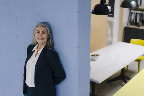 Porträt einer lächelnden grauhaarigen Geschäftsfrau vor einer blauen Wand in einem Loftbüro - GUSF03349
