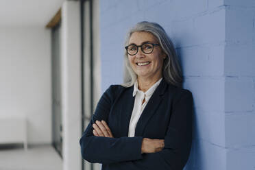 Portrait of smiling grey-haired businesswoman standing at a blue wall - GUSF03357