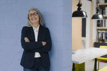 Porträt einer glücklichen grauhaarigen Geschäftsfrau vor einer blauen Wand in einem Loftbüro - GUSF03359