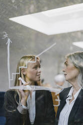 Ältere und junge Geschäftsfrau, die eine Tabelle auf einer Glasscheibe im Büro besprechen - GUSF03412