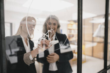 Mature and young businesswoman drawing chart on glass pane in office - GUSF03414