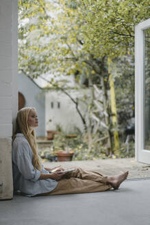 Junge Frau sitzt an der Terrassentür und benutzt ein Tablet - GUSF03419