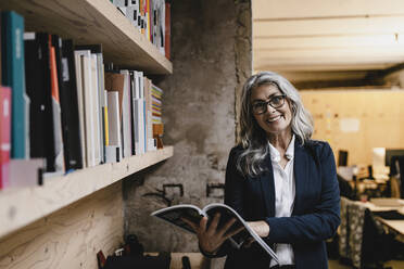 Portrait of smiling grey-haired businesswoman holding a magazine in a loft office - GUSF03452