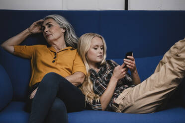 Woman using smartphone on couch next to her mother - GUSF03466