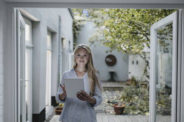 Portrait of smiling young woman standing at terrace door using tablet - GUSF03482