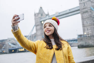 London, Vereinigtes Königreich, Mädchen mit Wollmütze und gelbem Mantel macht ein Selfie vor der Tower Bridge - DGOF00495