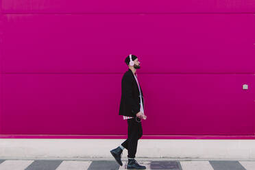 Junger Mann mit Kopfhörern und Smartphone geht an einer rosa Wand entlang - ERRF02783