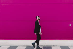 Young man with headphones and smartphone walking along a pink wall - ERRF02783