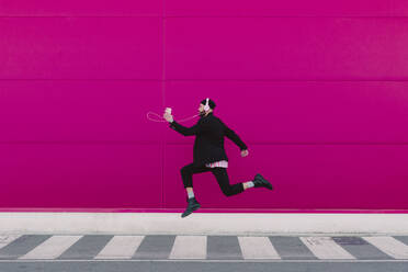 Junger Mann hört Musik und springt vor einer rosa Wand - ERRF02788