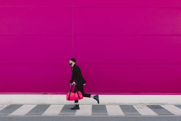 Young man with travelling bag running in front of a pink wall - ERRF02792