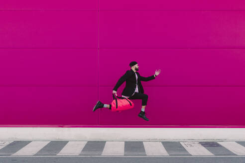 Young man jumping with travelling bag in front of a pink wall - ERRF02793