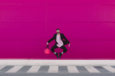 Junger Mann springt mit Reisetasche vor einer rosa Wand, lizenzfreies Stockfoto