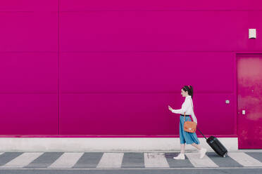 Young woman using smartphone and walking with trolley along a pink wall - ERRF02808