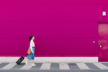 Young woman using smartphone and walking with trolley along a pink wall - ERRF02809