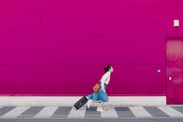 Junge Frau mit Smartphone läuft mit Trolley entlang einer rosa Wand - ERRF02810