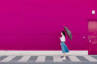 Junge Frau mit Regenschirm vor einer rosa Wand - ERRF02812