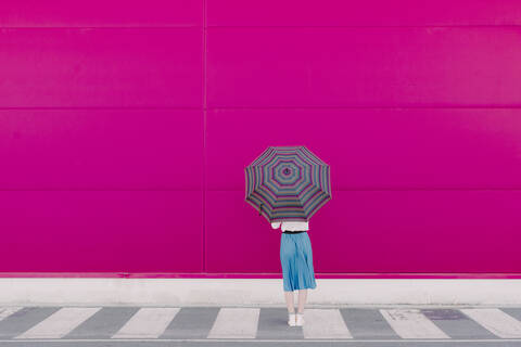 Rückansicht einer jungen Frau mit Regenschirm, die vor einer rosa Wand steht, lizenzfreies Stockfoto