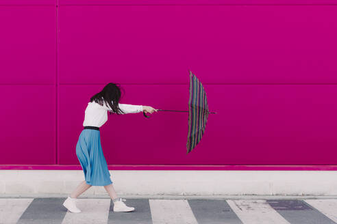 Junge Frau hält Regenschirm vor einer rosa Wand - ERRF02817