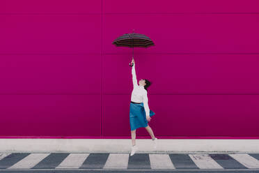 Junge Frau springt mit Regenschirm vor einer rosa Wand - ERRF02819