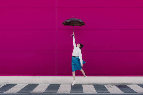 Young woman jumping with umbrella in front of a pink wall - ERRF02819