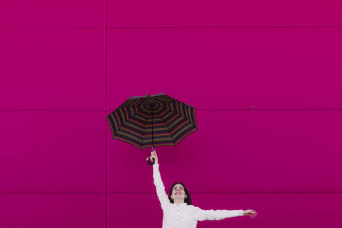 Young woman jumping with umbrella in front of a pink wall - ERRF02820
