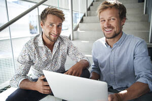 Porträt von zwei lächelnden jungen Geschäftsleuten, die mit einem Laptop auf einer Treppe sitzen - PNEF02368