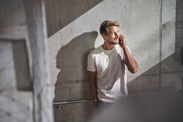 Junger Mann mit T-Shirt telefoniert an einer Betonwand - PNEF02384