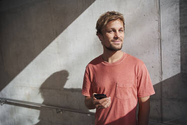 Porträt eines Mannes mit rotem T-Shirt, der sein Handy an eine Betonwand hält - PNEF02390