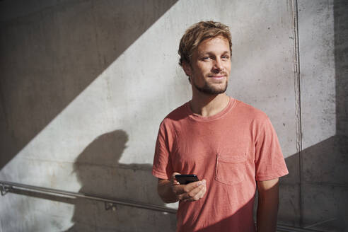 Porträt eines Mannes mit rotem T-Shirt, der sein Handy an eine Betonwand hält - PNEF02390