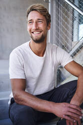 Porträt eines lächelnden jungen Mannes mit weißem T-Shirt, der auf einer Treppe sitzt - PNEF02403