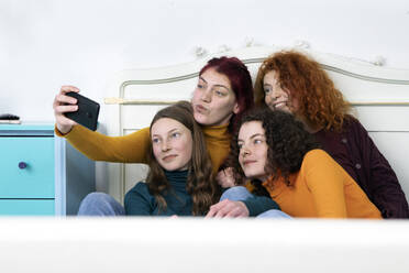 Mother and her three daughters sitting together on bed taking selfie with smartphone - PSTF00666