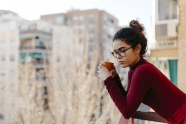 Portrait of young woman wearing red turtleneck pullover, holding a cup and standing on a balcony - TCEF00220