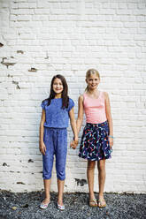 Portrait of two smiling girls standing hand in hand at a wall - SODF00683