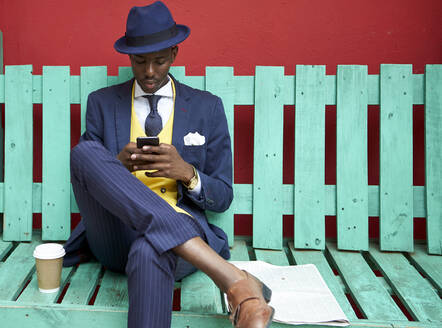 Young businessman wearing old-fashioned suit and hat sitting on a green bench checking his phone - VEGF01606