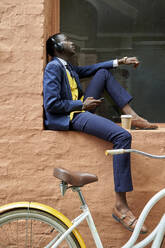 Stilvoller junger Geschäftsmann mit Fahrrad und altmodischem Anzug sitzt auf einem Fenstersims und hört Musik über seine Kopfhörer - VEGF01622