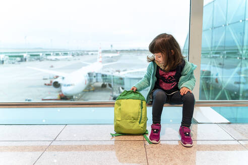 Mädchen mit Rucksack sitzt auf dem Flughafen vor einem Flugzeug - GEMF03467
